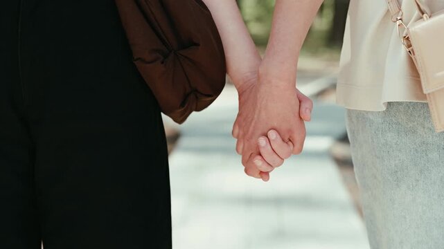Two people hold hands while walking through a park. They are on a pathway with trees on either side, enjoying their time together on a sunny day