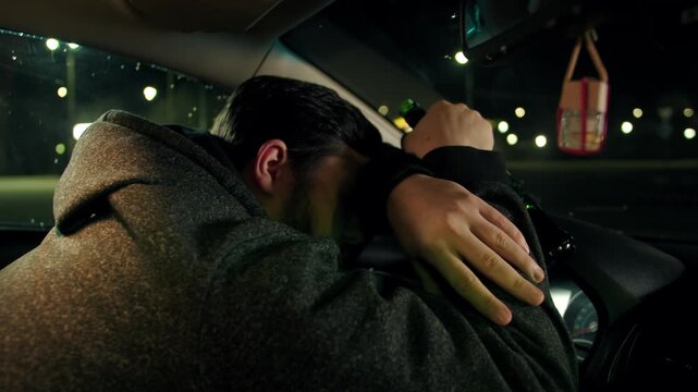 Asleep drunk adult man on parking. The driver folds his hands on the steering wheel, holds a bottle of alcohol and laid his head inside the car at night. The problem of alcohol addiction while driving