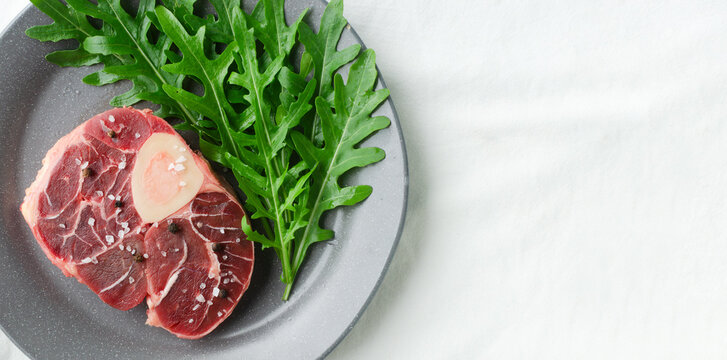 Osso buco, Cross section of raw beef shank with bone marrow with fresh arugula on a gray plate with a fork.