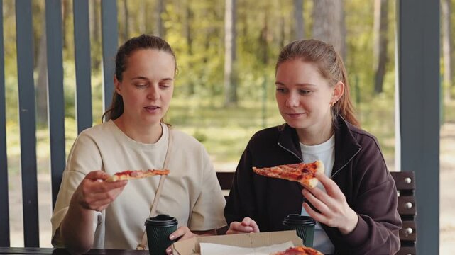 Two friends sit at a table in a park enjoying slices of pizza and warm drinks while having a friendly conversation on a sunny afternoon