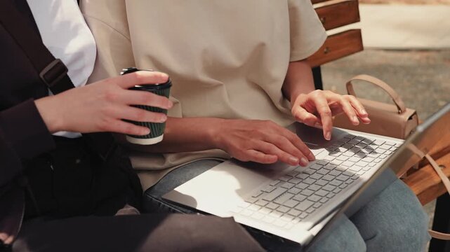Two individuals sit closely on a bench in a park. One holds a coffee cup while the other types on a laptop. They seem focused on their task