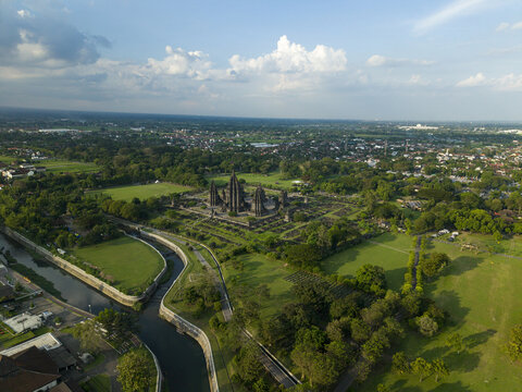 Aerial view of the ancient Prambanan Temple complex surrounded by lush green parkland and a winding canal under a cloudy sky in Kecamatan Prambanan, Daerah Istimewa Yogya, Indonesia.
