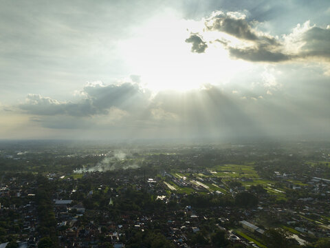 Aerial view of dramatic sun rays breaking through clouds over a rural landscape and residential area in Kecamatan Prambanan, Daerah Istimewa Yogya, Indonesia.