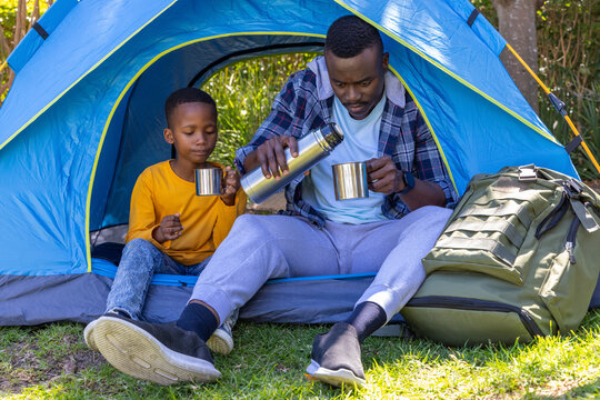 African American father and son sitting at blue dome tent entrance pouring thermos into metal cups