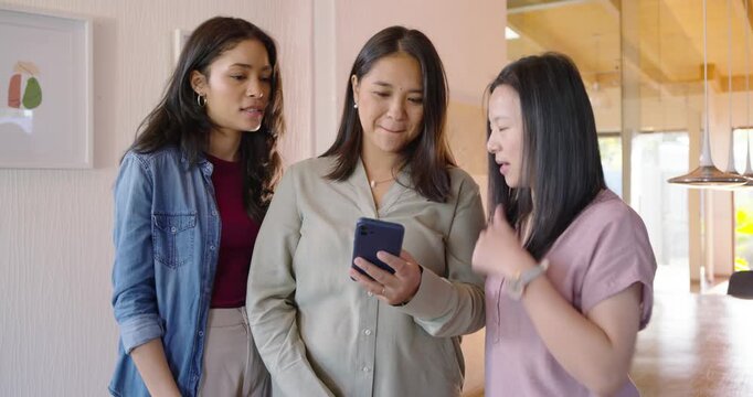 Diverse female friends watching center woman scrolling smartphone to view shared message at home