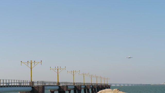 Distant view of a large passenger airplane approaching for landing over the sea. Commercial widebody jetliner flying above the water and yellow approach lighting system on the pier.