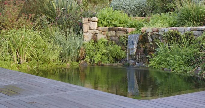 Camera panning right up, moving stone wall to center while spout pouring into pond creating ripples