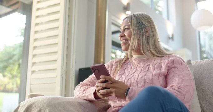 African American woman in pink sweater holding pink phone smiling scrolling after message on sofa