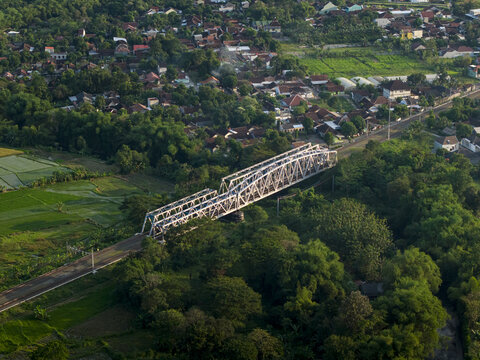 Aerial view of a large white steel truss bridge spanning over lush green trees and a river near a residential village Kecamatan Prambanan, Daerah Istimewa Yogya, Indonesia.