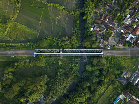 Aerial view of a railway bridge crossing a river surrounded by lush green rice fields and village houses in Kecamatan Prambanan, Daerah Istimewa Yogya, Indonesia.