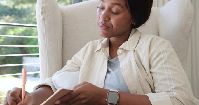 African American woman resting chin then lifting pen by window in armchair, journaling in notebook