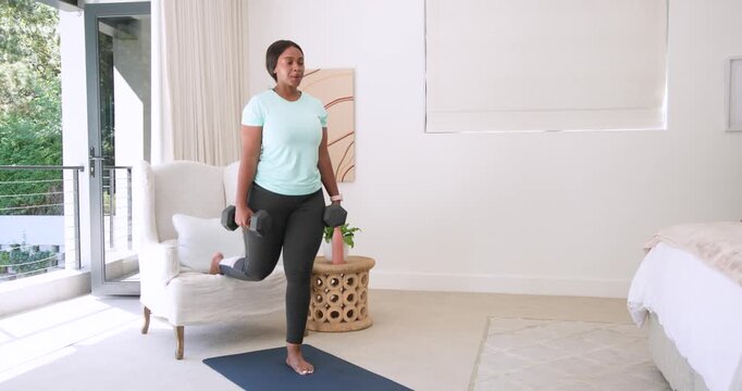 Adult African woman holding hex dumbbells, doing split squats using armchair on mat for strength