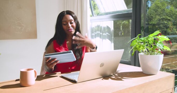 African American woman seeing tablet, pointing at laptop during call at desk wearing red blouse