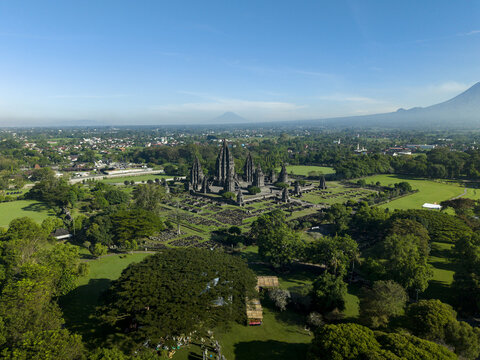 Aerial view of Prambanan Temple complex surrounded by lush green trees and a distant mountain under a clear blue sky in Kecamatan Prambanan, Daerah Istimewa Yogya, Indonesia.