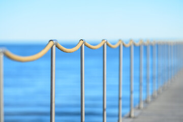 Obraz premium Long perspective of rope fence on pier with repeating metal posts and soft blue sea background.