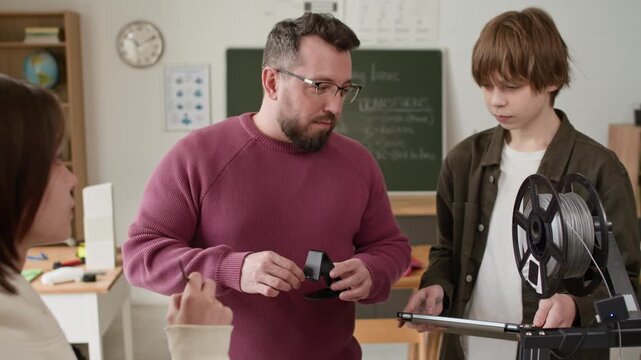 Male teacher demonstrating 3D printer plastic phone holder to interested students, gathered around 3D printer in modern classroom