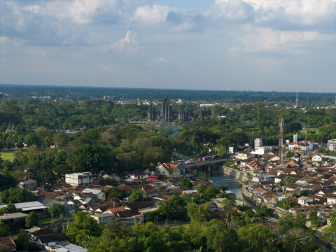Aerial view of Prambanan Temple and the surrounding lush green forest and residential houses under a cloudy sky in Prambanan, Yogyakarta, Indonesia.