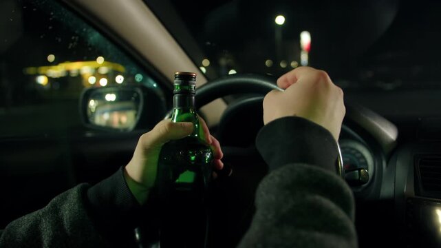 A closeup of a steering wheel and unscrewing the cap of a green bottle of alcohol. A man holds his hand on the steering wheel and drinks whiskey inside the car at night. Drinking alcohol while driving