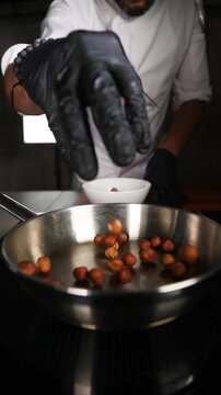 Chef echando un pu&ntilde;ado de avellanas en una sart&eacute;n de acero inoxidable. Un chef profesional a&ntilde;ade avellanas a una sart&eacute;n de acero. Proceso de tostado, preparaci&oacute;n de una receta gourmet artesanal.