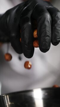 Chef echando un pu&ntilde;ado de avellanas en una sart&eacute;n de acero inoxidable. Un chef profesional a&ntilde;ade avellanas a una sart&eacute;n de acero. Proceso de tostado, preparaci&oacute;n de una receta gourmet artesanal.