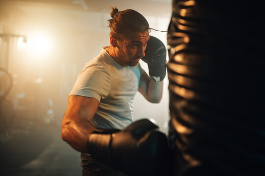Focused man exercising with boxing gloves on a heavy bag in a dimly lit fitness studio, improving health and stamina