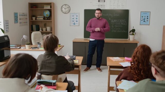 Handheld zoom out shot of male teacher standing near chalkboard with notes and giving lecture to group of attentive middle aged students in modern lassroom