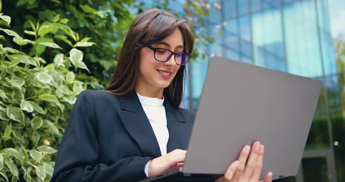 Portrait of beautiful entrepreneur woman carrying a laptop, working and typing message while sitting on bench at glass building background.