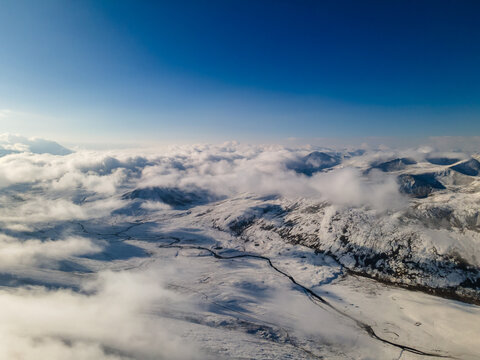 Aerial view of Babusar Pass with snow-covered mountain peaks, thick white clouds, and a winding road under a clear blue sky in Naran, KPK, Pakistan.