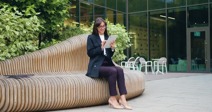 Young business woman with long hair sitting on a bench outside a modern office building, typing on a tablet and smiling at her work results