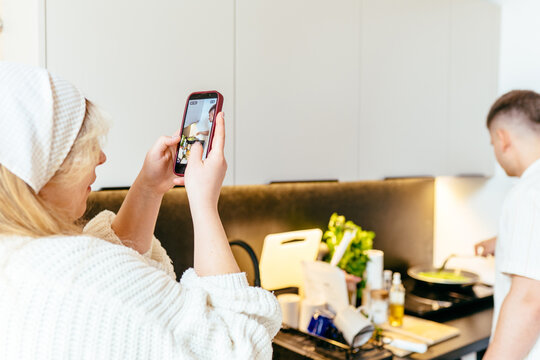 Young Caucasian woman recording partner cooking breakfast in Scandinavian tiny house kitchen. Digital lifestyle, social media content creation, cozy morning routine and modern couple life.