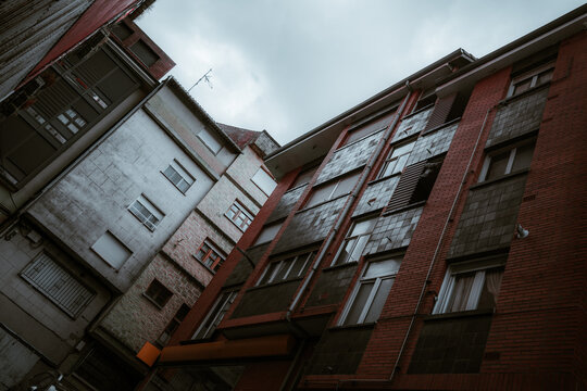Moody upward view inside a narrow old apartment courtyard, with red brick walls, shuttered windows, worn facades and cloudy sky creating a dark urban atmosphere
