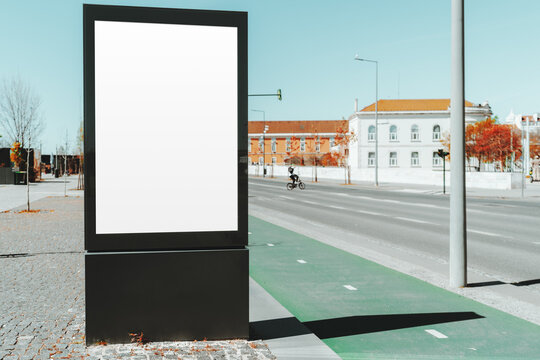 Large blank city advertising panel beside a green bike lane, with a silhouette of a cyclist crossing the street and bright buildings in the sunny urban defocused background and a copy space area