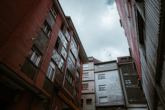 Looking upward in a tight residential courtyard framed by red brick and pale old facades, with shuttered windows, cloudy sky and muted low key ambience