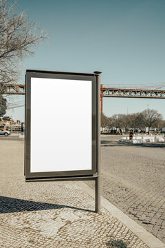 Empty vertical advertising panel on a sunny paved city square, ready for poster design display, with bridge structure, bare trees and urban traffic in soft background