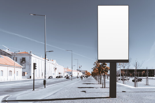 Blank vertical outdoor billboard on a wide sunlit avenue, ready for brand artwork, with white historic buildings, streetlights, trees and open sidewalk space; with a copy-space area on the left