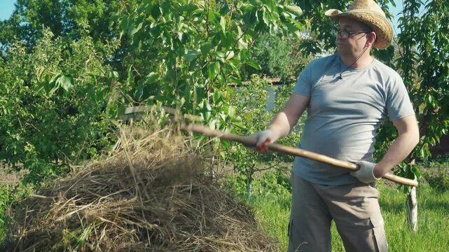 Gardener in a straw hat and gray shirt lifts hay with a pitchfork from a large pile in a sunny garden, surrounded by lush green trees and vibrant grass in the background