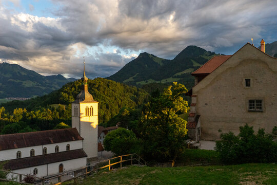 Gruyeres, Switzerland - 14 May 2026: View of the Church of Saint-Theodule and Gruyeres Castle against a backdrop of the Prealps mountains at sunset.