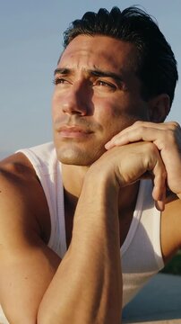 Vertical video: Leaning man in white tank top tilting toward sunlight on pool coping, posing