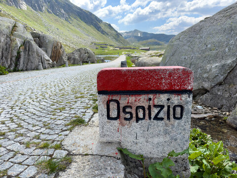 Gotthard Pass, Switzerland - 16 May 2026: View of a stone marker with the word Ospizio on a cobblestone road leading through the rugged alpine landscape of the Gotthard Pass.
