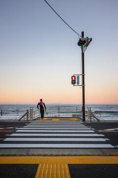 View of a surfer carrying a surfboard across a pedestrian crossing towards the ocean under a soft sunset sky Kamakura, Kanagawa, Japan.