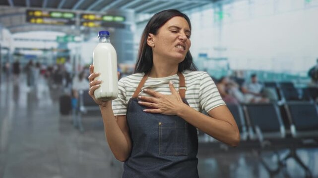 Young woman wearing apron holding a milk bottle with hand on chest, grimacing amid airport terminal seating and signage; disgust food safety.