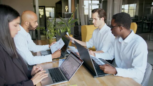 A diverse group sits in an office working with laptops. They analyze trading data to figure out the next steps for their development strategy. Collaboration is key in their discussions.