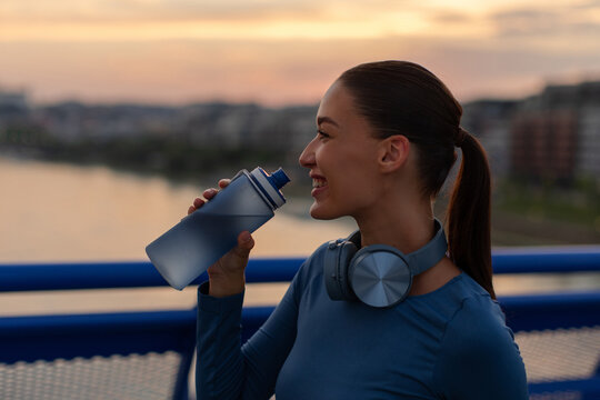 Young caucasian female athlete drinking water during outdoor fitness training at sunset