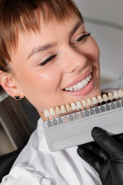 a dentist in black gloves holds a full tooth shade guide close to a smiling woman showing her bright white teeth