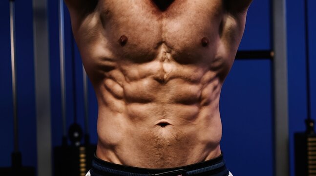 Muscular man shows six pack abs in gym. Fit male bodybuilder with defined abdominal muscles poses against blue background. Strength training, fitness, bodybuilding.