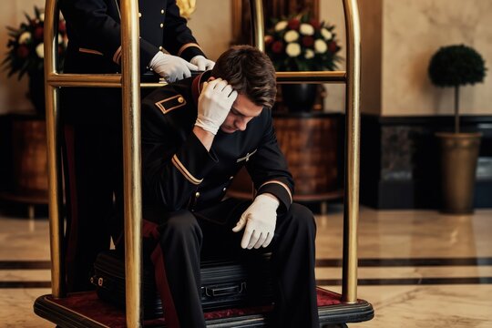 Tired doorman in uniform sits on luggage cart with hand on head. Another staff member stands nearby. Hotel lobby busy with guests needing baggage assistance. Exhausted worker endures long shift.