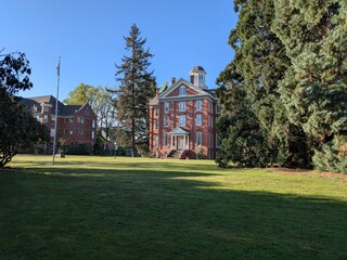 Obraz premium : Historic collegiate campus with green lawns, sculpture, flagpole, and classic brick academic buildings beneath a vibrant spring sky.