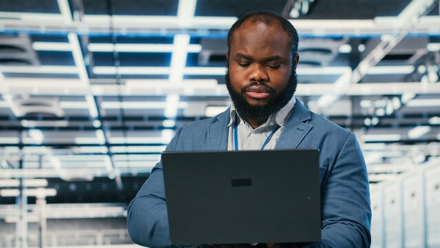 Software developer in server farm using laptop, doing network management virtualization, African American man in data center overseeing IT infrastructure supercomputers with notebook, camera B.