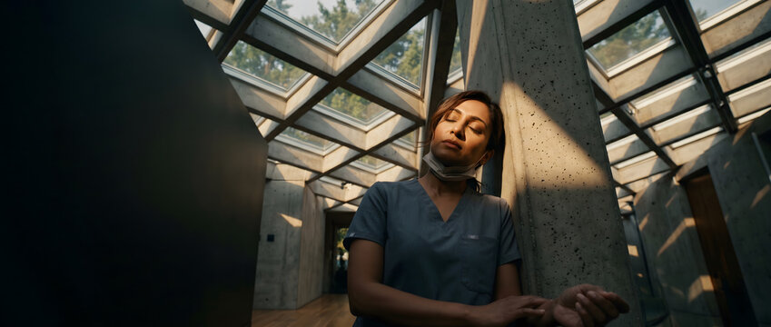 Tired female doctor resting against concrete pillar in modern hospital building, exhausted healthcare worker in blue scrubs taking a peaceful mental health break under skylight, burnout concept.