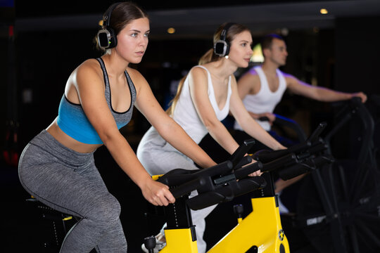 Girl in earphone spend time in gym, improve physical shape with exercises. Female sport studio visitor listen to music and uses exercise bike to pump leg muscles. Strength and motivation, fitness goal